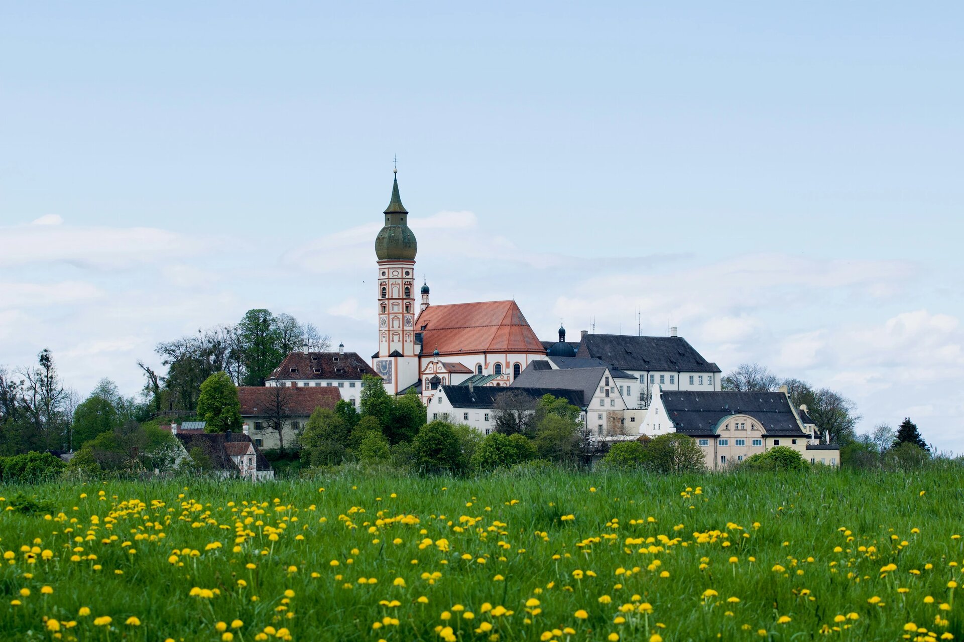 A scenic view of the historic Andechs Abbey (Kloster Andechs) in Bavaria, Germany. The monastery sits atop a gentle hill, overlooking a lush green meadow dotted with bright yellow dandelions under a soft, partly cloudy sky.