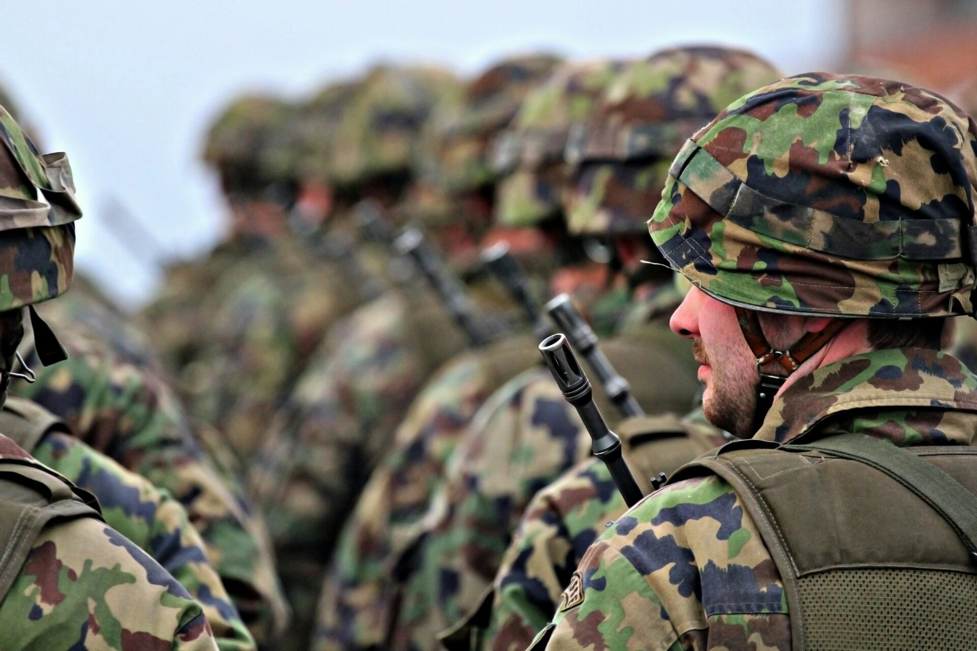 A striking side-profile perspective captures a column of Swiss soldiers standing in a precise formation. The shallow depth of field creates a beautiful bokeh effect, blurring the long line of troops while keeping the foreground soldier in sharp, detailed focus. He wears a camouflage-covered helmet and a tactical vest, with the barrel of an assault rifle pointing upward. The lighting is soft and diffuse, characteristic of a cloudy day, which enhances the rich greens and browns of the woodland camo. The image evokes a strong sense of readiness, discipline, and the quiet intensity of military ser