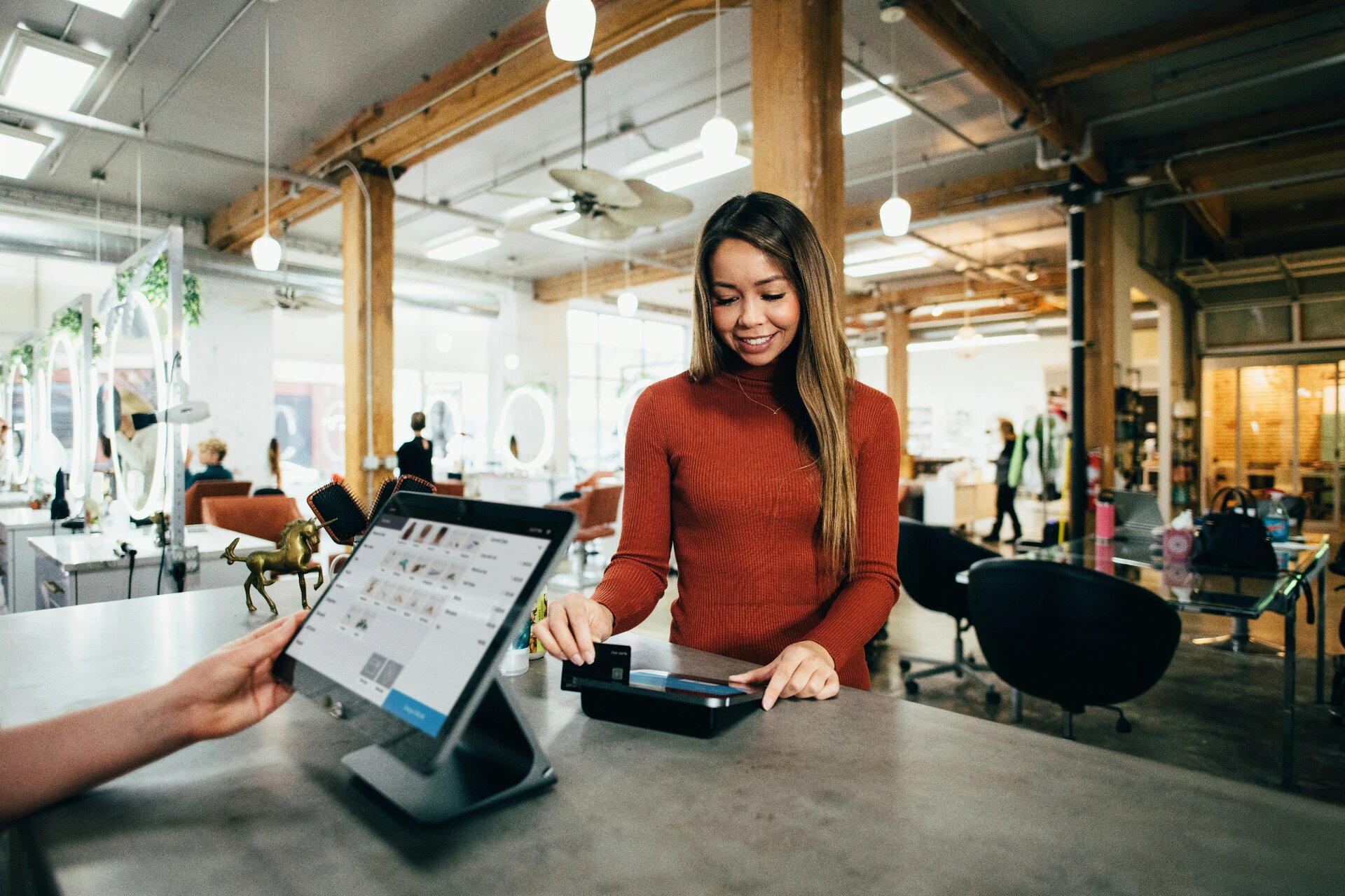 Smiling woman makes contactless payment at modern retail POS terminal with tablet register system. Industrial-style store interior showcases contemporary cashless shopping experience.