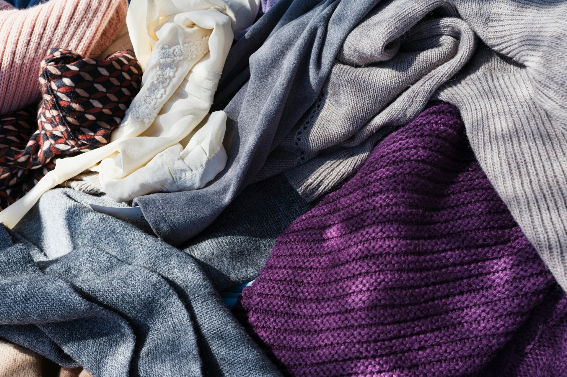 Close Up of Colorful Knitted Sweaters Laying on Top of Each Other - A close-up of a pile of colorful knitted sweaters, including a pink sweater, a white sweater, a grey sweater, a dark blue sweater, and a purple sweater.